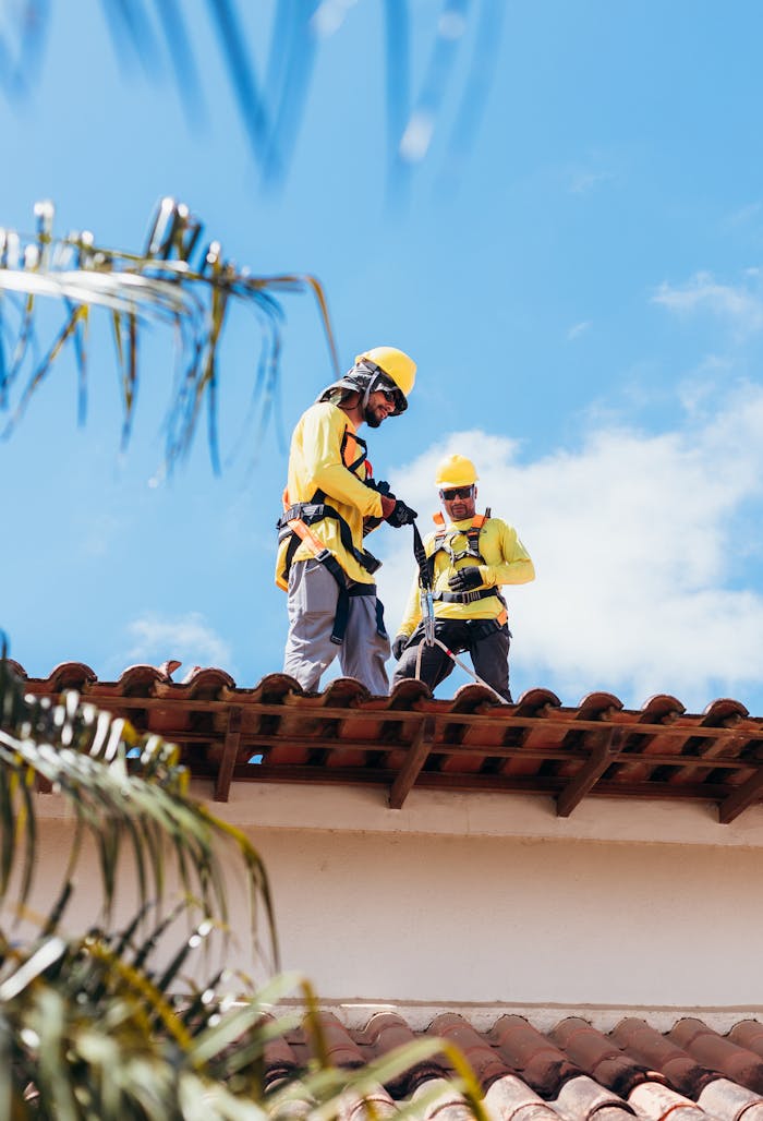 Two construction workers in safety gear working on a tiled roof under a clear blue sky.
