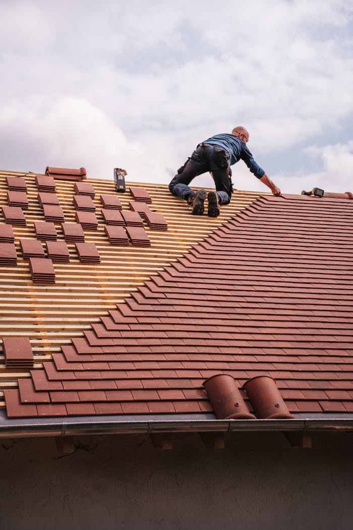 A roofer carefully installs shingles on a new residential roof under a cloudy sky.